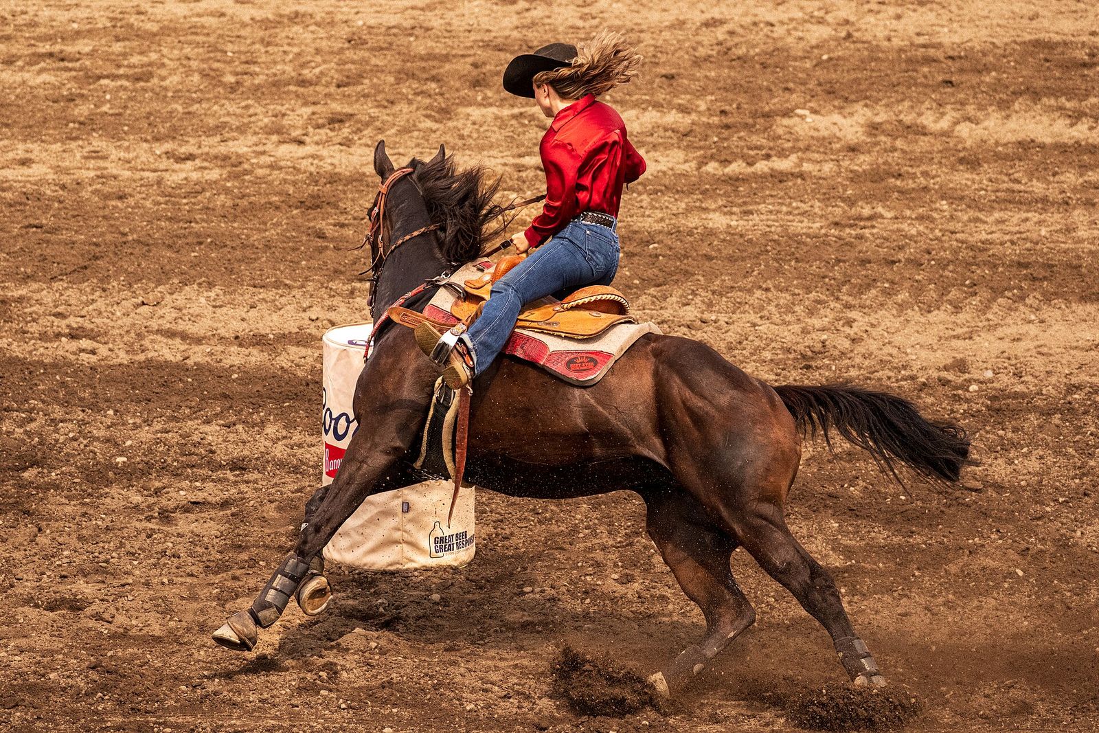A rider in a red shirt and cowboy hat rounding a barrel on a galloping horse in a dirt arena